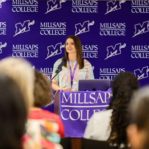 Young woman standing at a podium with a Millsaps College banner on it, speaking to a crowd.