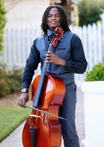 Young, African-American man dressed in a black shirt, grey vest and pants and a bowtie playing the chello.