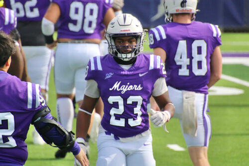 Young African-American man in a football uniform, purple jersey with the white number 23 and white pants, running on the field.