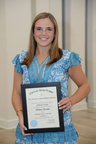 A young woman with straight blonde hair, wearing a light blue dress holds a black-framed certificate.