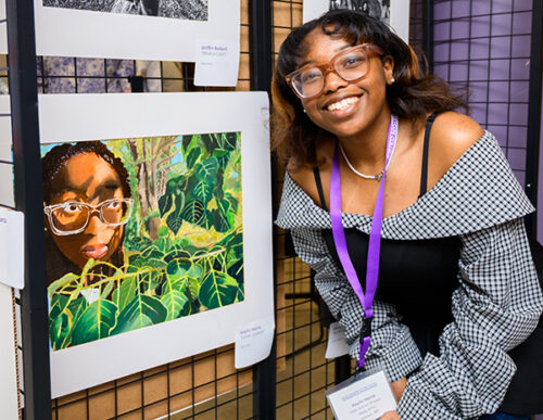 Artist Kaylin Harris poses with her artwork featuring an African-American girl with glasses on peaking out of green foliage, almost jungle-looking.