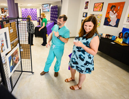 A man in surgical scrubs and a woman in a blue dress with black shapes on it, smile as they look at the art on exhibit.